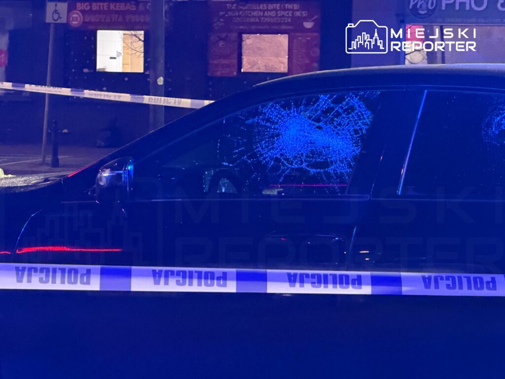 Czarny samochód osobowy z rozbitą szybą, otoczony taśmą policyjną, w tle widoczne zamknięte lokale.