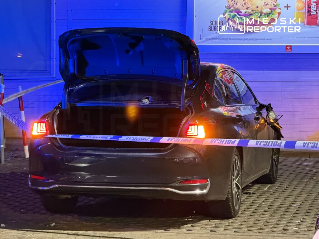 Czarny samochód z otwartą klapą bagażnika, otoczony taśmą policyjną, zaparkowany w oświetlonym miejscu.