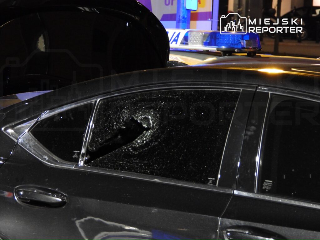 Rozbita szyba w czarnym samochodzie osobowym, obok policyjny radiowóz z włączonymi sygnałami świetlnymi.