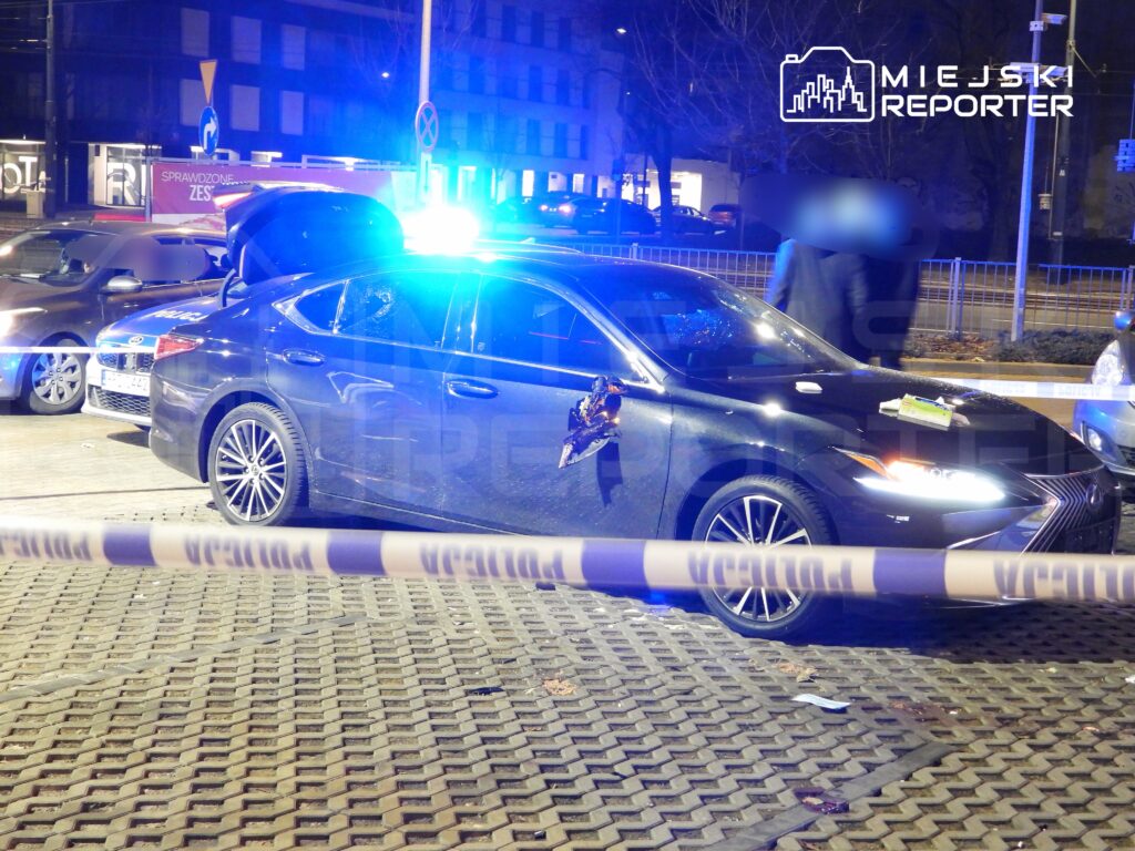 Czarny samochód osobowy z otwartą pokrywą bagażnika i włączonymi światłami policyjnymi na ulicy, otoczony taśmą zabezpieczającą.