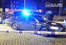 Czarny samochód osobowy z otwartą pokrywą bagażnika i włączonymi światłami policyjnymi na ulicy, otoczony taśmą zabezpieczającą.