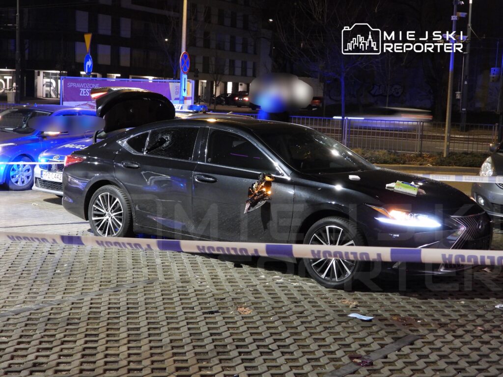Czarny samochód osobowy z uszkodzoną szybą stoi w miejscu zdarzenia, otwarta klapa bagażnika, w tle radiowóz policyjny.
