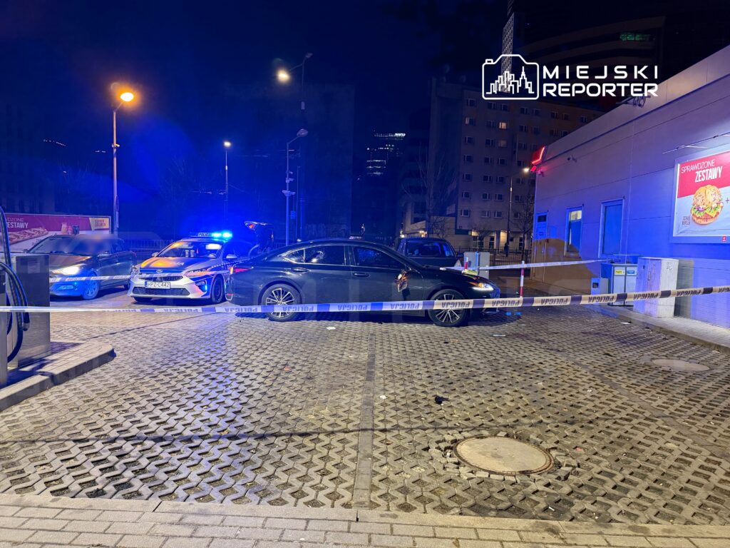 Policyjna akcja w nocy, z radiowozem zaparkowanym obok zablokowanej drogi, oświetlonej niebieskimi światłami.