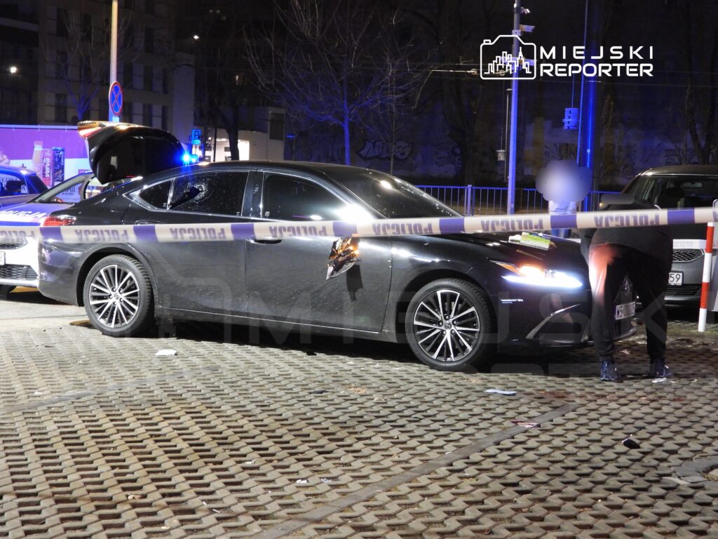 Czarny samochód osobowy z uszkodzoną szybą stoi w obszarze oznaczonym taśmą policyjną. Obok, funkcjonariusz bada okoliczności zdarzenia.
