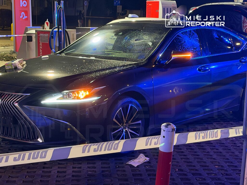 Czarny samochód osobowy z uszkodzoną szybą stoi na stacji benzynowej, oświetlony niebieskimi światłami policyjnymi.
