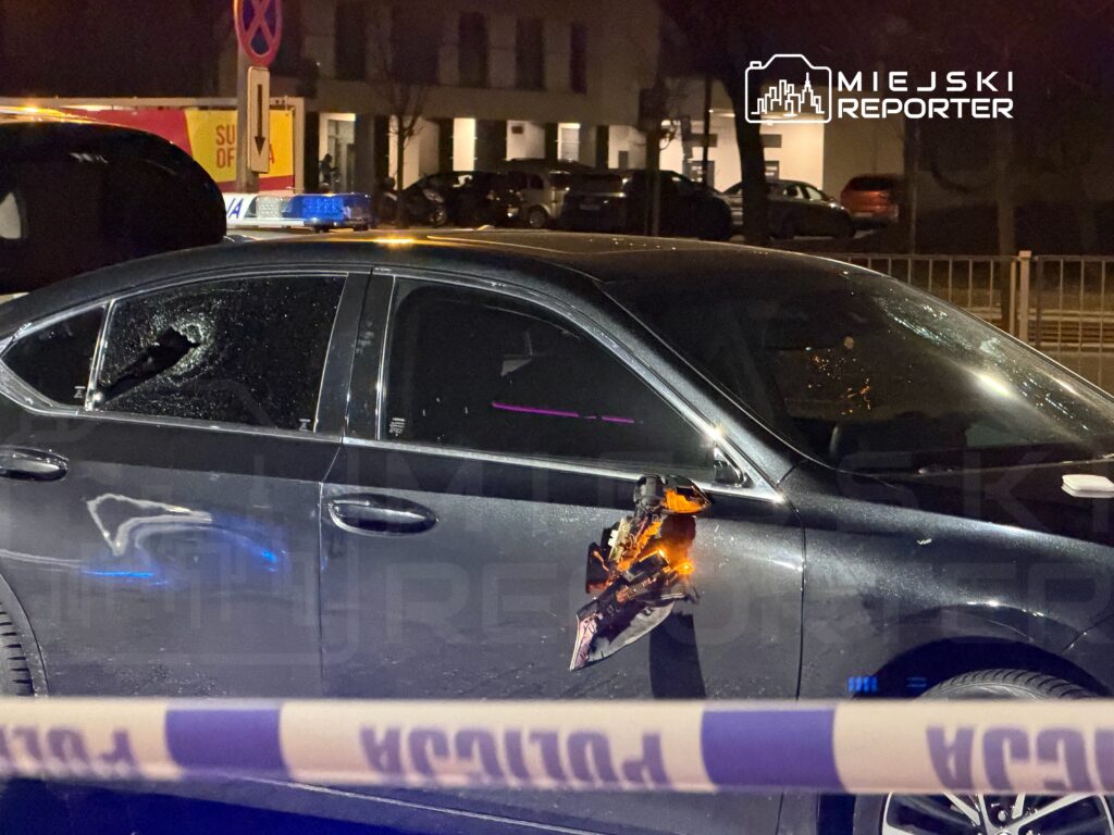 Czarny samochód osobowy z wybitym oknem i uszkodzeniami karoserii, otoczony taśmą policyjną w nocy.