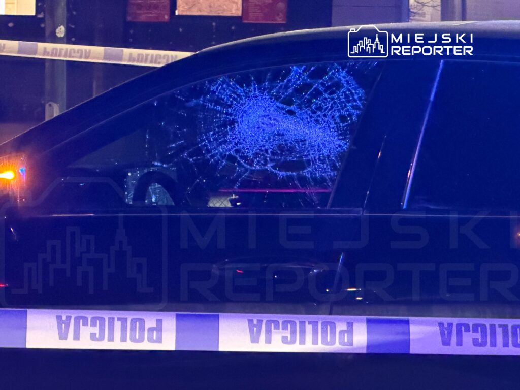 Uszkodzone okno samochodu z pęknięciami, otoczone taśmą policyjną w nocy. W tle widać nieostre światła miasta.