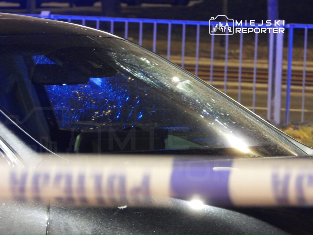 Czarny samochód osobowy z pękniętą szybą, oświetlony niebieskimi światłami policyjnymi, znajduje się za taśmą zabezpieczającą.