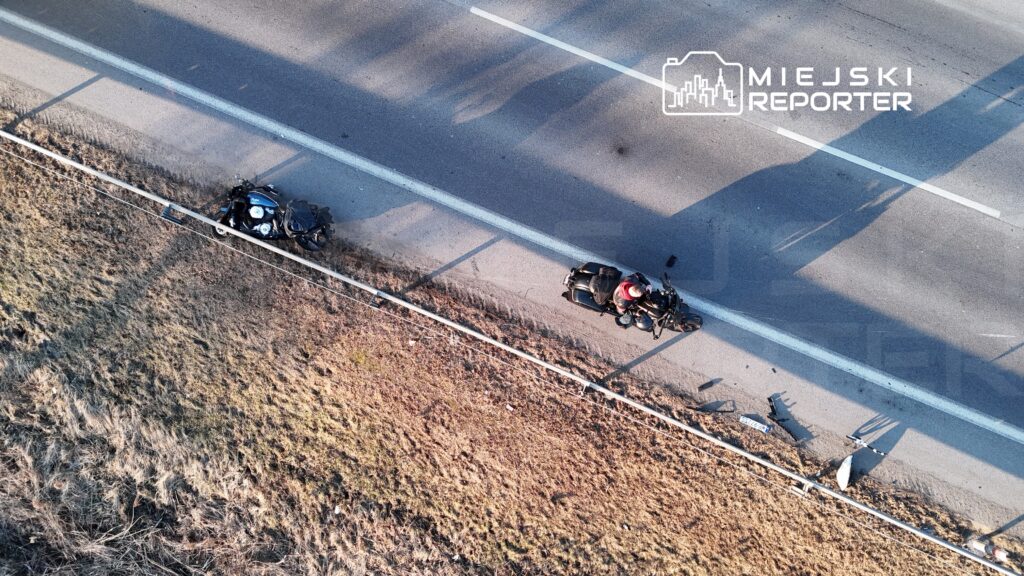 Motocykl leży na poboczu drogi, obok stoi motocyklista w czerwonej kurtce, a na jezdni leżą fragmenty pojazdu.