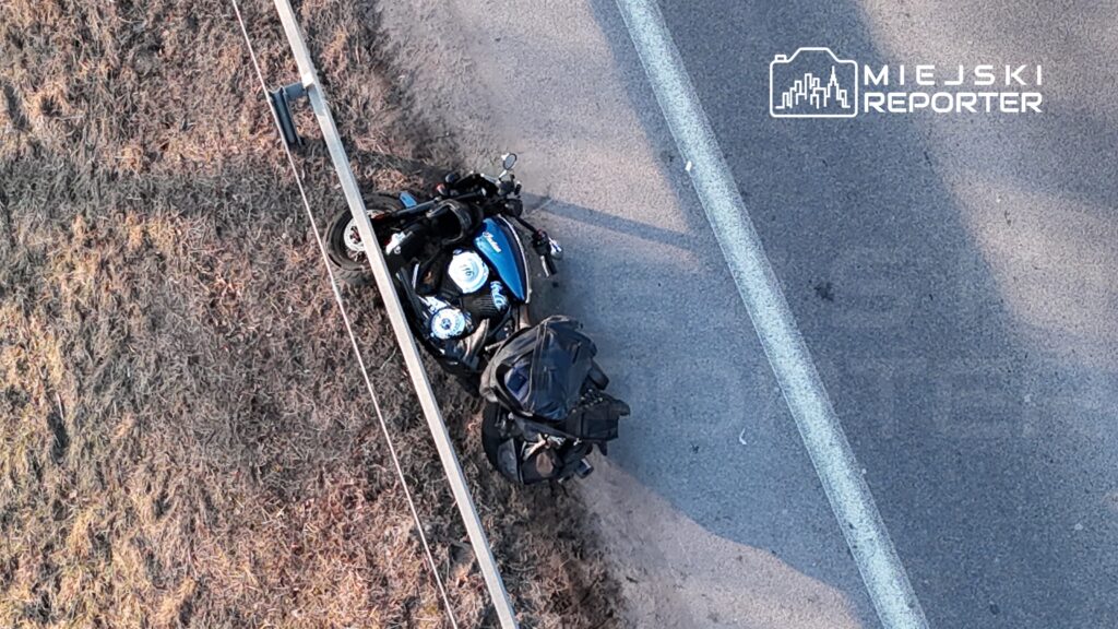 Motocykl leży na poboczu drogi, obok metalowej barierki, z torbą na bagażniku. W tle widoczna asfaltowa nawierzchnia.