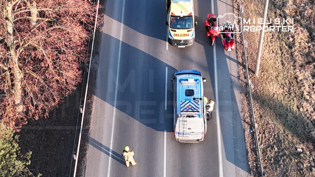 Na drodze stoi radiowóz policyjny, obok dwóch funkcjonariuszy rozmawiających z osobą w mundurze. W tle widać karetkę pogotowia.