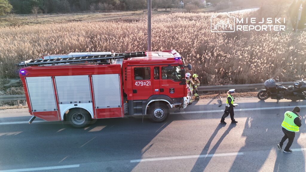 Czerwony wóz strażacki zatrzymany przy drodze, obok strażacy pomagają poszkodowanemu motocykliście, w tle trawa.