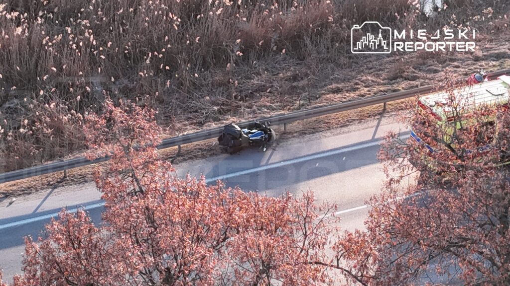 Na poboczu drogi leży motocykl obok bariery ochronnej, a w tle widać pojazd służb ratunkowych.