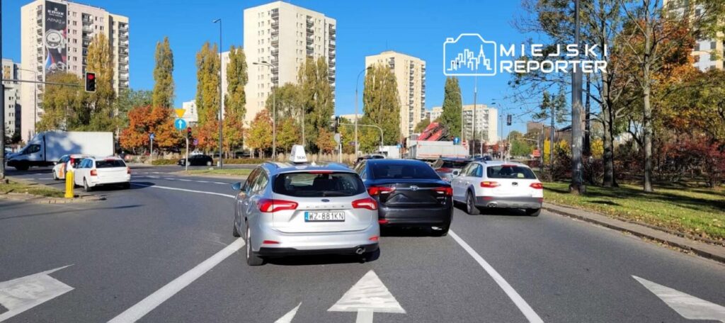 Ruch samochodowy na rondzie z widokiem na wysokie bloki mieszkalne i drzewa w jesiennych kolorach.