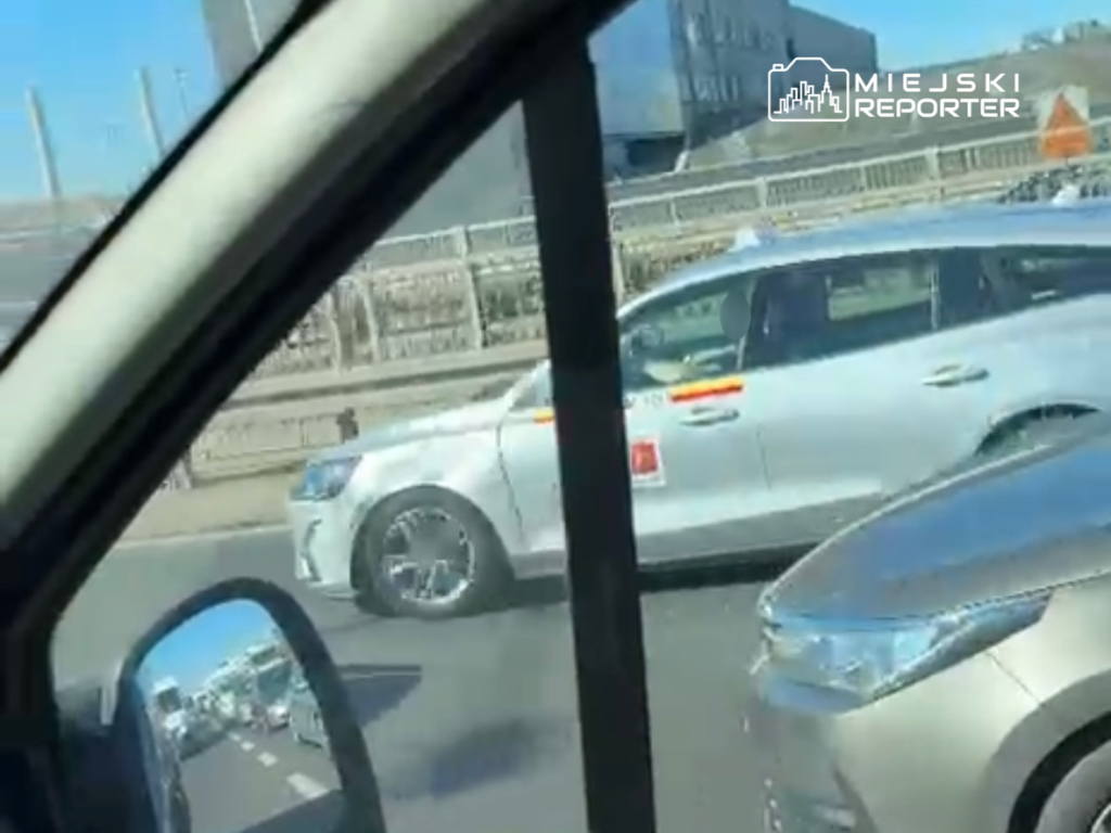 Biały samochód osobowy z widoczną flagą na drzwiach, poruszający się w ruchu drogowym.