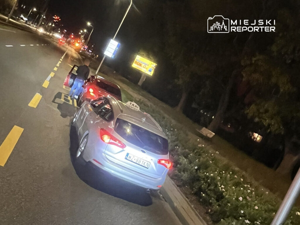 Mężczyzna w ciemnej odzieży otwiera drzwi czerwonego samochodu na ulicy w nocy, obok taksówki.