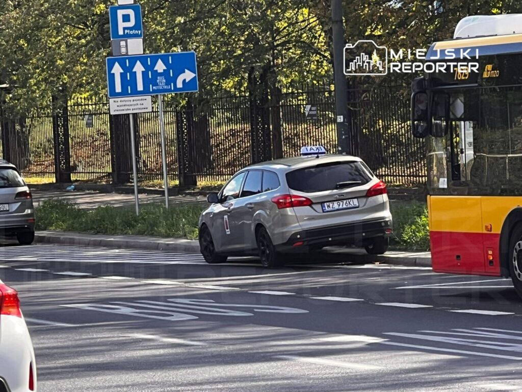 Taksi zaparkowane na pasie ruchu obok przystanku autobusowego, z widoczną sygnalizacją drogową.