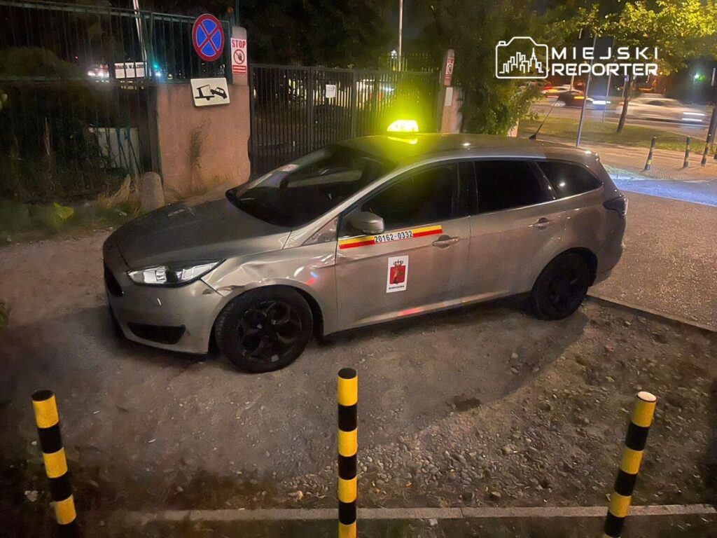 Szary samochód z oznaczeniem policyjnym zaparkowany na chodniku obok znaku zakazu parkowania.