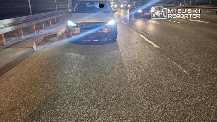 Uszkodzony samochód zaparkowany na jezdni w nocy, w otoczeniu innych pojazdów poruszających się w tle.