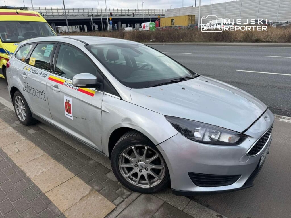 Srebrne taxi z napisem "Airport" zaparkowane przy drodze, obok karetki pogotowia.