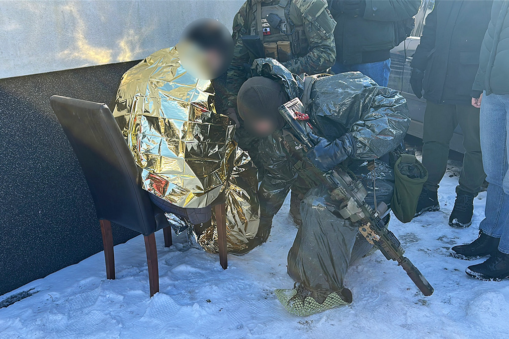 Żołnierz w maskowaniu sprawdza osobę owiniętą w folię termiczną na śniegu, wokół grupka ludzi w odzieży zimowej.