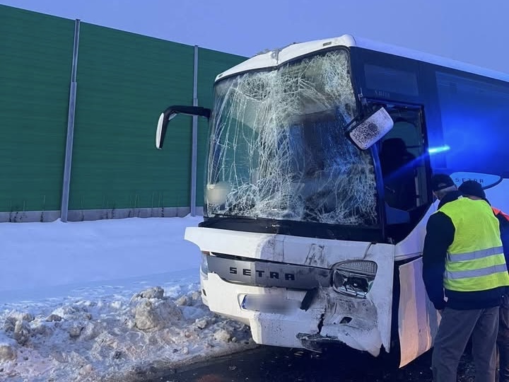 Autobus marki Setra z rozbitą szybą i uszkodzonym przodem stoi na zaśnieżonej drodze, obok pracownik w kamizelce odblaskowej.