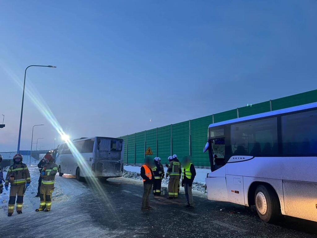 Grupa strażaków i ratowników medycznych przy uszkodzonym autobusie na ośnieżonej drodze, w tle zielony ekran dźwiękochłonny.