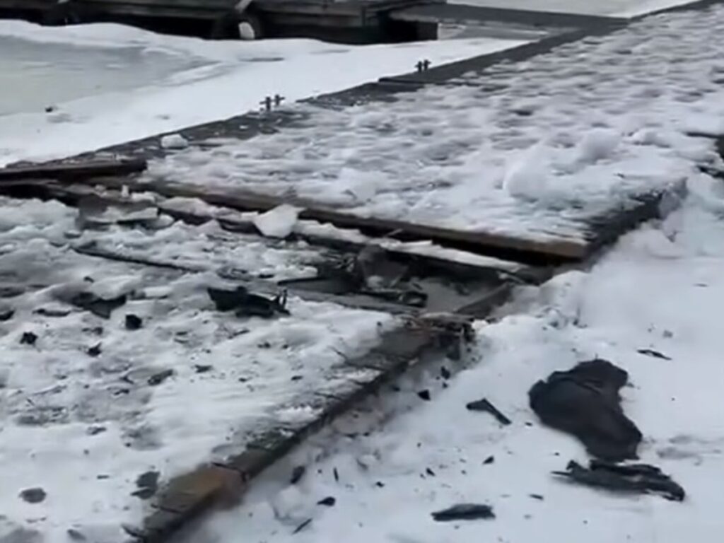 Zniszczona drewniana platforma pokryta śniegiem i lodem, z fragmentami desek leżącymi obok.