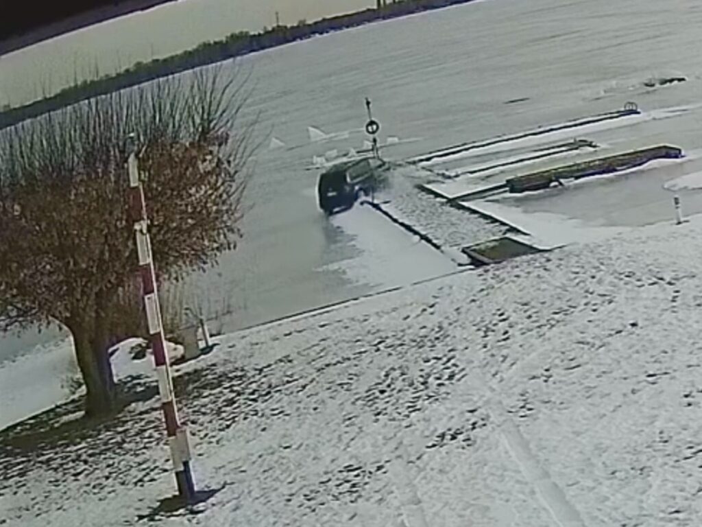 Samochód wjeżdża na zaśnieżoną rampę wodną, obok widoczne słupki i zamarznięta woda.