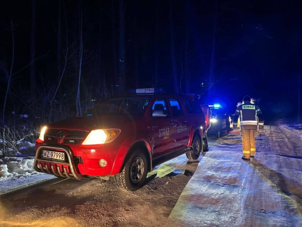 Czerwony wóz strażacki stoi na oświetlonej drodze, a strażak w mundurze zmierza w kierunku pojazdu.