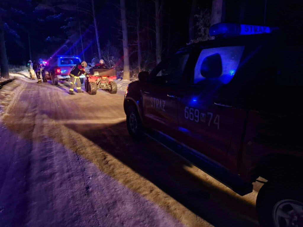 Strażacy w mundurach pracują przy wciąganiu quada na drogę w lesie, w nocy, z użyciem policyjnych świateł.