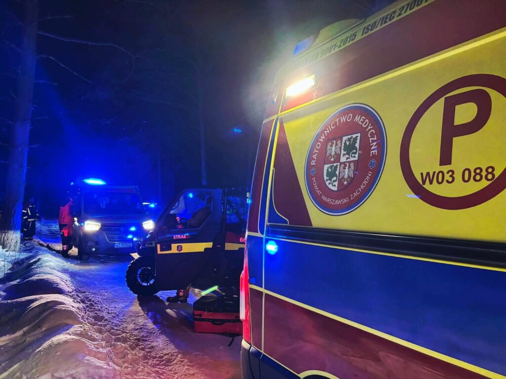 Karetka pogotowia i pojazd straży pożarnej w akcji ratunkowej na ośnieżonej drodze w lesie, w nocy, z niebieskimi światłami.