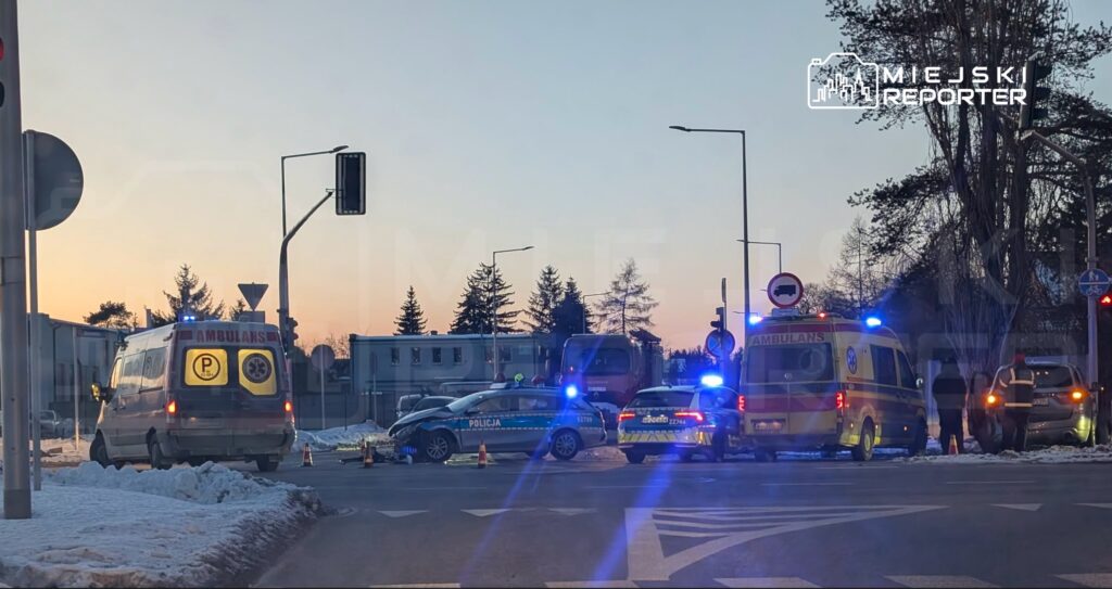 Na skrzyżowaniu stoją dwa ambulanse oraz radiowóz policyjny, a w tle widać zarys budynków i drzewa.