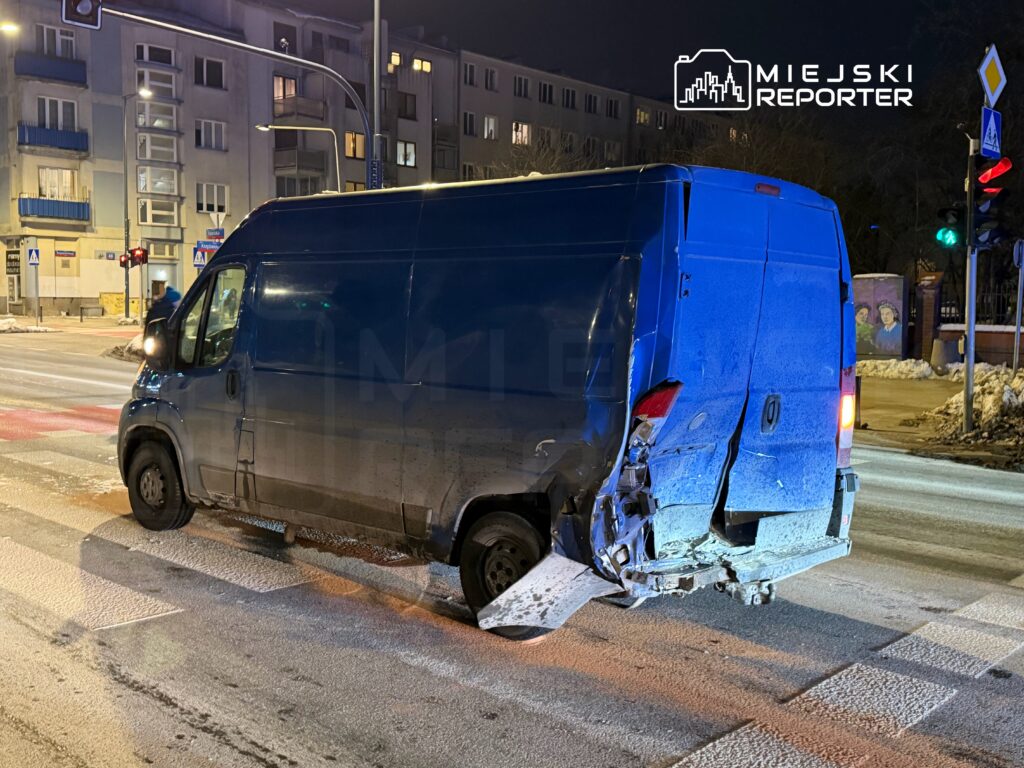 Niebieski furgon z uszkodzeniem tyłu stoi na skrzyżowaniu, obok przejścia dla pieszych, w nocy.