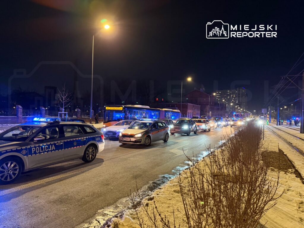 Policyjny radiowóz z niebieskimi światłami blokuje ruch na ulicy w nocy, obok stoją inne samochody w korku.