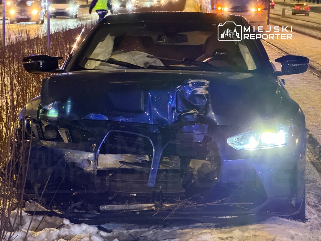 Uszkodzony samochód osobowy zaparkowany na poboczu drogi, z widocznymi uszkodzeniami przodu i włączonymi światłami.