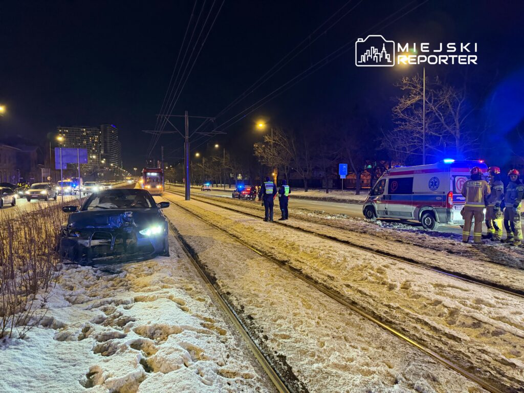 Uszkodzony samochód osobowy stoi na poboczu, obok grupy ratowników i policji w nocy, w oświetlonym mieście.