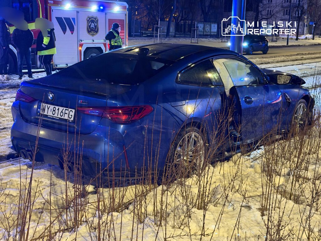 Niebieskie BMW M4 zaparkowane na poboczu, obok grupy ludzi i wozu strażackiego w tle.
