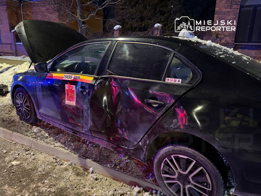 Uszkodzony samochód osobowy z otwartą maską stoi na zaśnieżonej ulicy, obok widoczne ślady kolizji.