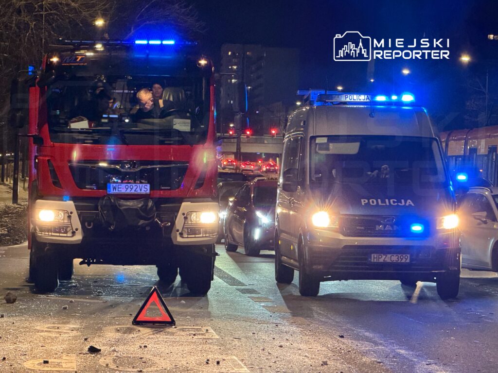 Czerwony wóz strażacki oraz policyjny furgon z włączonymi sygnałami świetlnymi stoją na drodze, wokół leżą drobne kamienie.