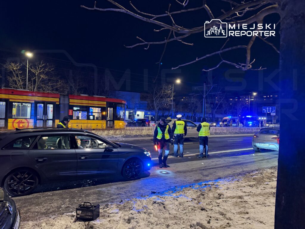 Grupa policjantów w odblaskowych kamizelkach prowadzi czynności na oświetlonej ulicy, obok zatrzymanego samochodu.