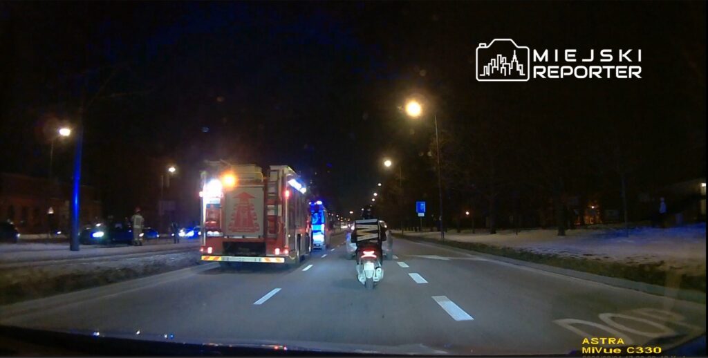 Na drodze nocą stoi straż pożarna z włączonymi sygnałami świetlnymi, a obok przejeżdża skuter dostawczy.