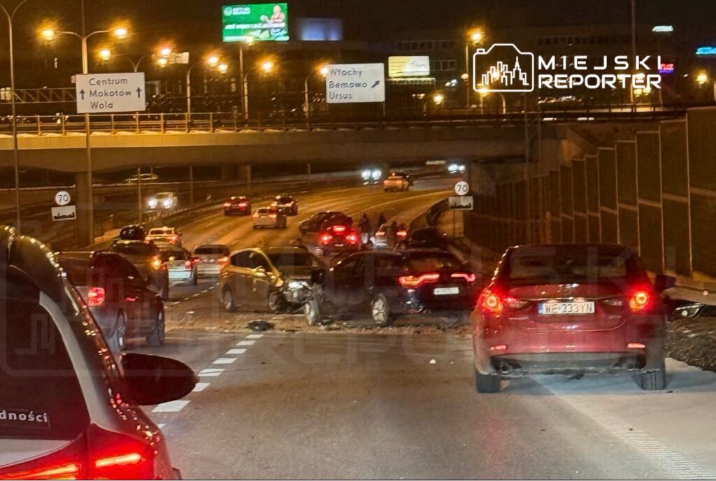 W nocy na ruchliwej drodze widać zderzenie kilku samochodów, a kierowcy stoją obok pojazdów.