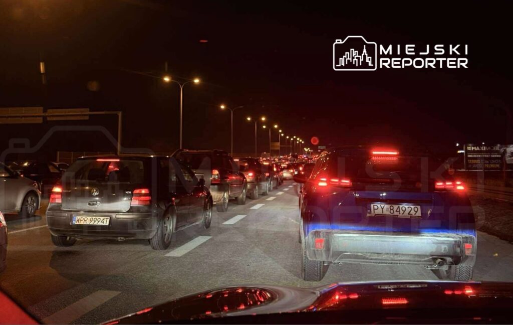 Samochody stoją w korku na ulicy w nocy, oświetlone przez latarnie i światła pojazdów.
