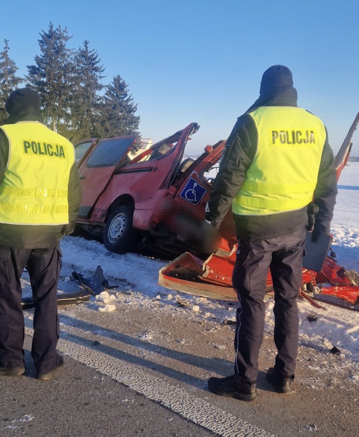 Dwóch policjantów w odblaskowych kamizelkach bada uszkodzony czerwony pojazd zaparkowany na zaśnieżonej drodze.