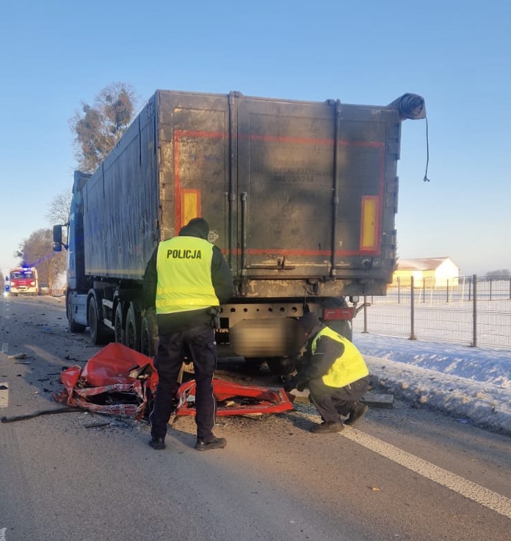 Dwóch policjantów w odblaskowych kamizelkach bada wrak czerwonemu samochodu osobowego na drodze obok ciężarówki.
