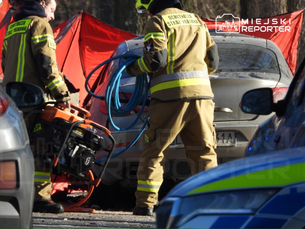 Strażacy w mundurach z narzędziem hydraulicznym i wężem w rękach pracują przy uszkodzonym samochodzie na drodze.