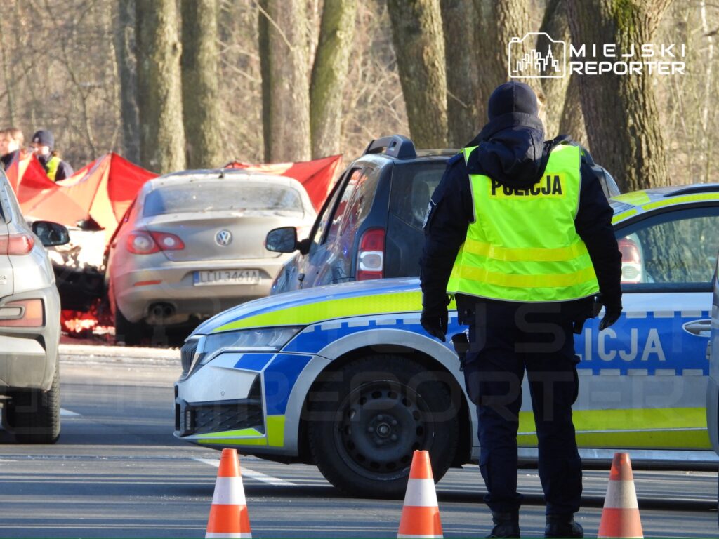 Policjant w kamizelce odblaskowej kieruje ruchem drogowym obok wypadku samochodowego, w tle czerwone namioty.