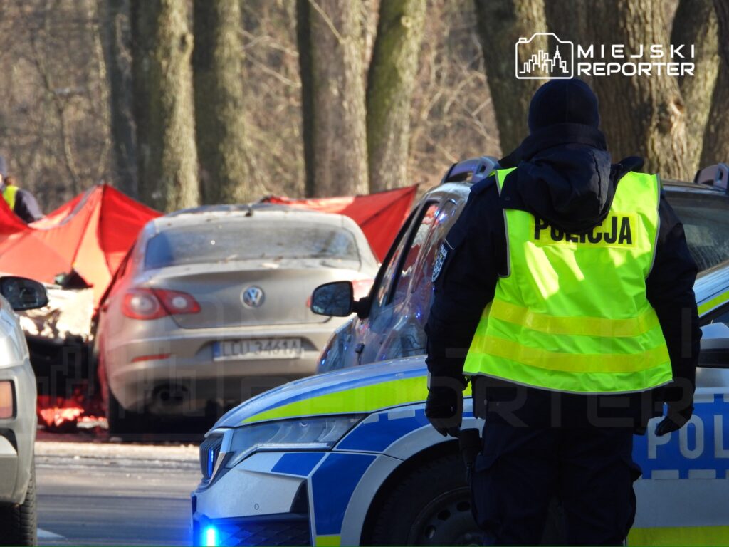 Policjant w kamizelce odblaskowej obserwuje miejsce zdarzenia z czerwonym namiotem oraz uszkodzonym samochodem.