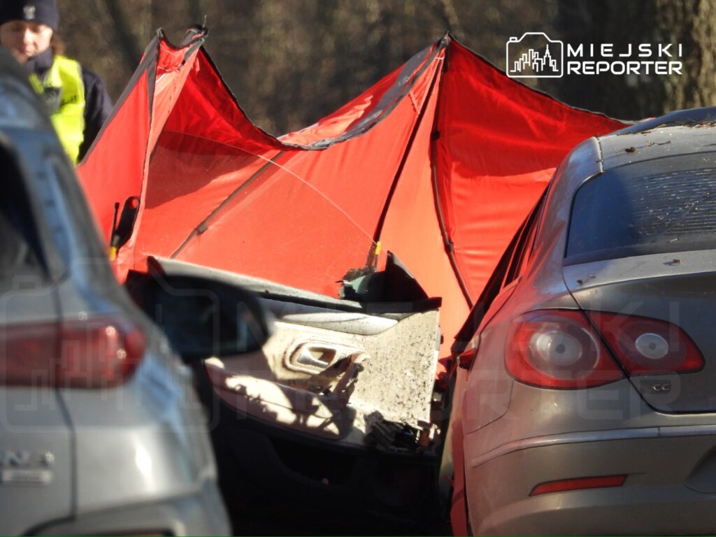 Czerwony namiot zabezpieczający stoi obok uszkodzonego samochodu, a w tle widać inne pojazdy. Policjant bada miejsce zdarzenia.